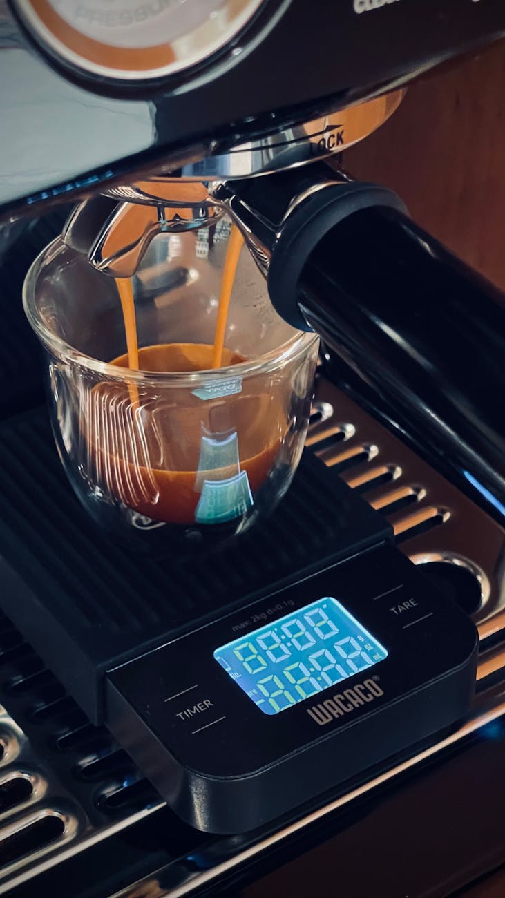 A compact premium coffee scale sitting on an espresso machine's drip tray, measuring an espresso shot being pulled, steam rising, focused on the scale's display showing weight and timer, high contrast, studio lighting, professional
