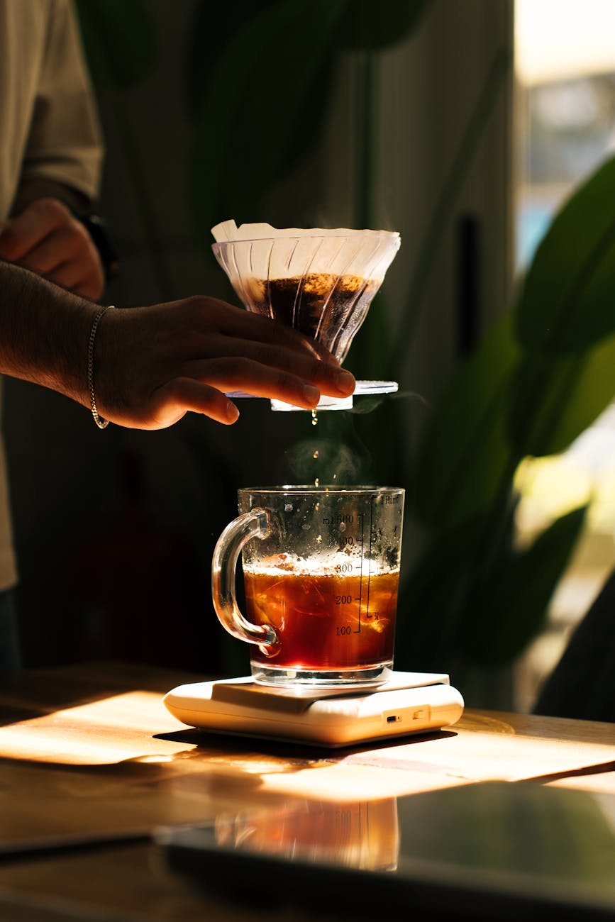 A close-up shot of hands carefully pouring hot water over ground coffee in a pour over dripper, steam rising, in a sunlit kitchen. The focus is on the intricate flow of water and the bloom of the coffee grounds, with a gooseneck kettle visible.
