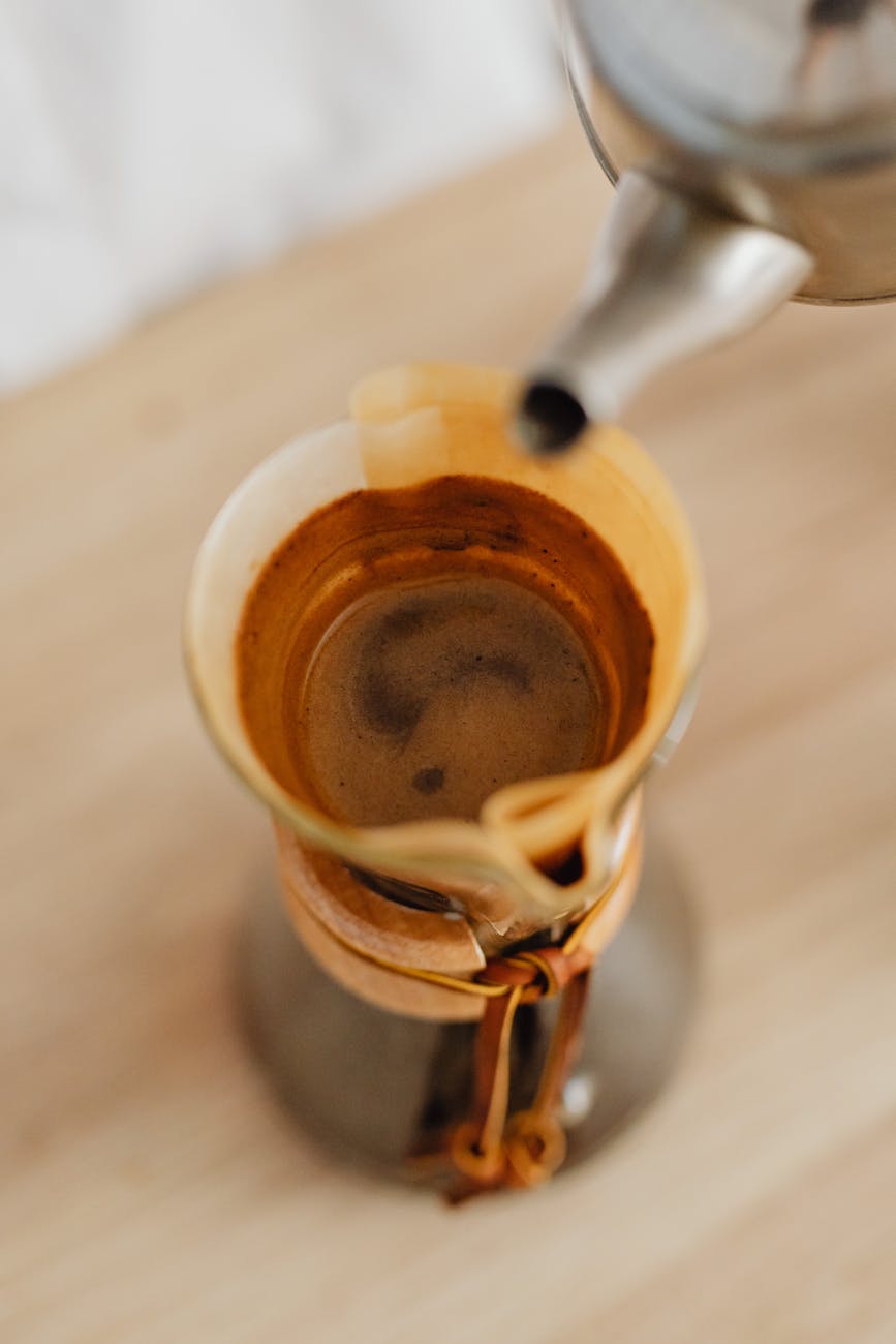 A top-down view of a pour over brewing setup on a wooden counter, showing a gooseneck kettle precisely pouring water, a coffee dripper with grounds, a digital scale, and a timer. The scene is well-lit and clean.